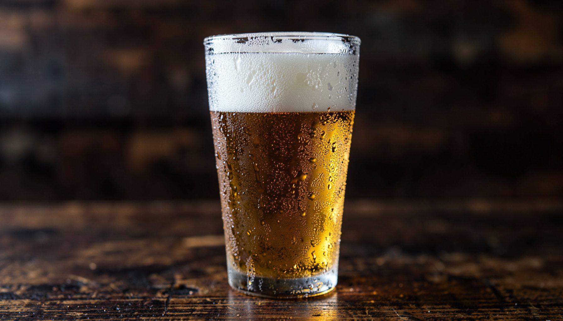 A tall glass of golden beer with a foamy white head, covered in condensation droplets, on a dark wooden surface